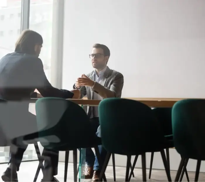 Due uomini siedono uno di fronte all'altro al tavolo di un ufficio e discutono.