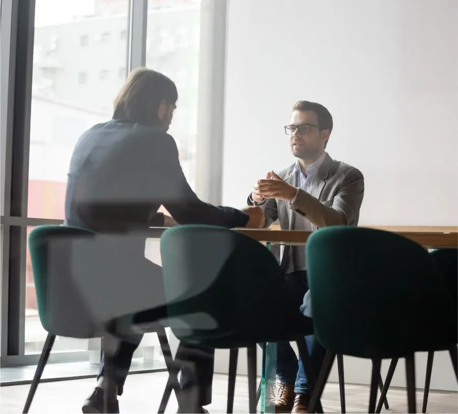 Due uomini sono seduti al tavolo di un ufficio e stanno conversando.